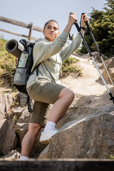 Low angle view of young short haired woman traveler with backpack holding trekking poles and looking away while standing near hill with stones, Translation of tattoo: love — Stock Photo