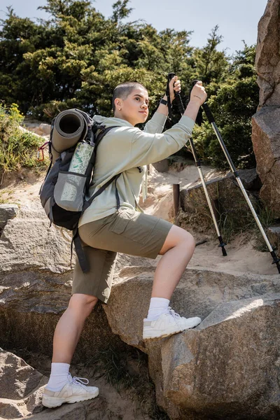 Short haired female traveler with backpack and map holding trekking poles while standing on stones near hill at background, tranquil hiker finding inner peace on trail, Translation of tattoo: love — Stock Photo