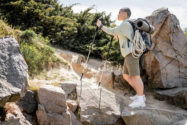 Side view of young short haired woman tourist with backpack holding trekking poles walking on hill with stones and grass at background, tranquil hiker finding inner peace on trail, summer — Stock Photo