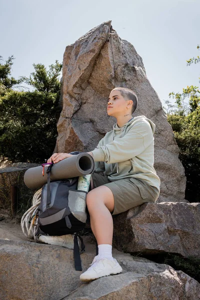 Молодая коротковолосая женщина в повседневной одежде сидит рядом с рюкзаком с картой на камне на холме с природой на заднем плане, спокойный турист находит внутренний мир на тропе, лето — стоковое фото