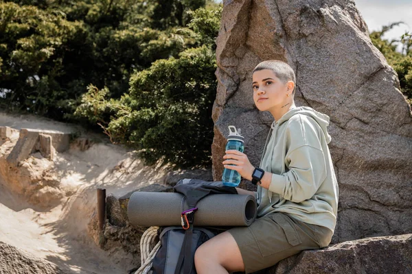 Молода короткошерста і татуйована жінка турист тримає спортивну пляшку, дивлячись і сидячи біля рюкзака на камені з природою на фоні, спокійний пішохід знаходить внутрішній спокій на стежці — стокове фото