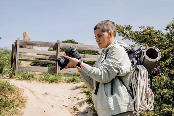 Молодая женщина путешественник с короткими волосами с повседневной одеждой и рюкзак с цифровой камерой, стоя с холмом и живописный пейзаж на заднем плане, турист находит вдохновение в природе — стоковое фото