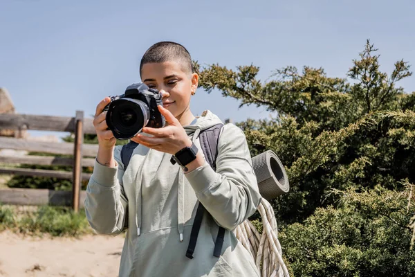 Positive junge, kurzhaarige und tätowierte Touristin mit Rucksack, die mit Digitalkamera fotografiert, während sie mit der Natur und blauem Himmel im Hintergrund steht, Wanderin, die Inspiration in der Natur findet — Stockfoto