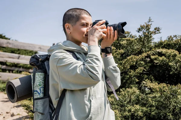 Young tattooed and short haired female tourist with backpack and map taking photo on digital camera while standing with nature at background, travel photographer, summer, Translation of tattoo: love — Stock Photo