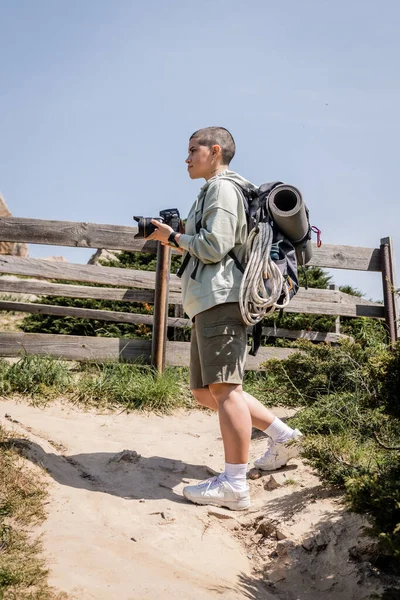 Seitenansicht einer jungen kurzhaarigen Wanderin mit Rucksack und Reiseausrüstung, die eine Digitalkamera in der Hand hält und auf einem Hügel mit Himmel im Hintergrund steht, Wanderin, die Inspiration in der Natur findet — Stockfoto