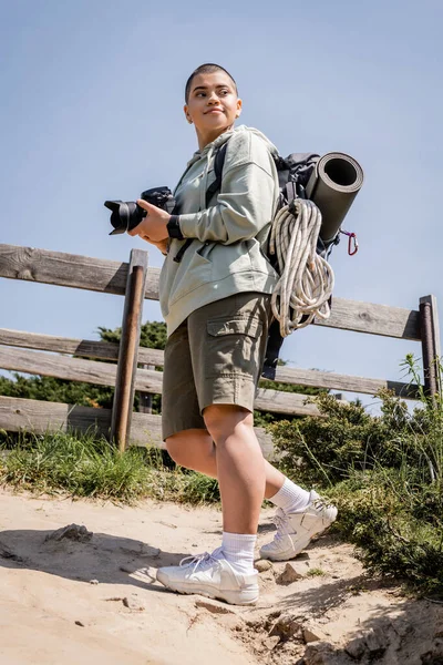 Low angle view of joyful young short haired female traveler with backpack and travel equipment holding digital camera and looking away while standing on hill, travel photographer — Stock Photo