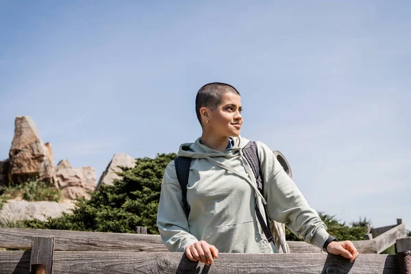 Joven y positiva excursionista femenina de pelo corto con mochila mirando hacia otro lado mientras está de pie cerca de una valla de madera con cielo azul al fondo, excursionista trekking a través del paisaje, verano - foto de stock