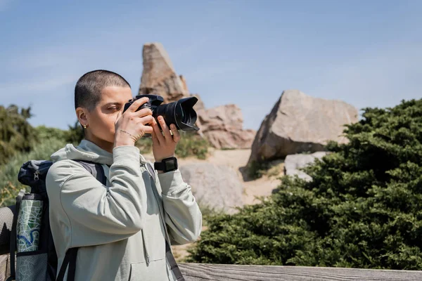Молодая коротковолосая и татуированная туристка с рюкзаком и картой, фотографирующая на цифровой камере, стоя на заднем плане с природой, Перевод татуировки: любовь — стоковое фото