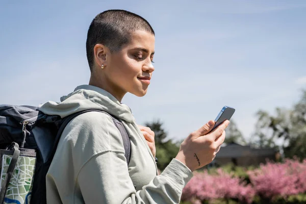 Young tattooed and short haired female tourist with map in backpack using smartphone while hiking with blurred nature and blue sky at background, exploring new horizons, Translation of tattoo: love — Stock Photo