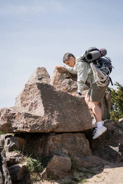 Seitenansicht einer jungen kurzhaarigen Touristin mit Rucksack und Reiseausrüstung, die auf Steine mit blauem Himmel und Natur im Hintergrund klettert, um neue Horizonte zu erkunden, Sommer — Stockfoto