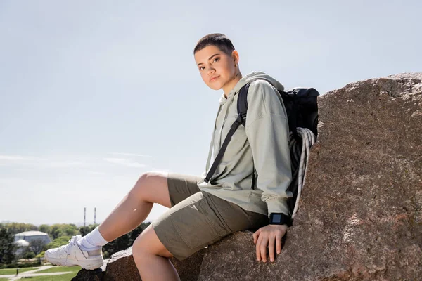 Junge kurzhaarige Wanderin mit Rucksack und Fitness-Tracker blickt in die Kamera und sitzt auf Steinen mit blauem Himmel und Natur im Hintergrund, lebendige Reiseerlebnisse, Sommer — Stockfoto