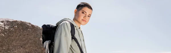 Young short haired and tattooed female tourist in casual clothes with backpack looking at camera while standing near stone with blue sky at background, vibrant travel experiences, banner — Stock Photo