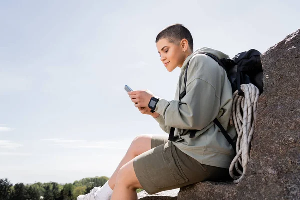 Молодая коротковолосая женщина-путешественница с рюкзаком и туристическим оборудованием, используя смартфон, сидя на камне с природой и голубым небом на заднем плане, живой опыт путешествия, лето — стоковое фото