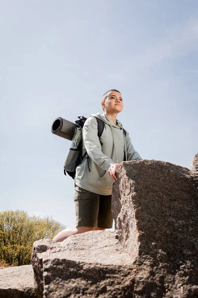 Junge kurzhaarige Wanderin in lässiger Kleidung mit Rucksack, Karte und Fitnessmatte, die wegschaut, während sie in der Nähe von Steinen mit blauem Himmel und Natur im Hintergrund steht, lebendige Reiseerlebnisse — Stockfoto