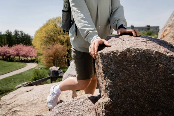 Cropped view of young female hiker with backpack and fitness tracker standing near stones on hill with blurred nature at background, vibrant travel experiences, summer — Stock Photo
