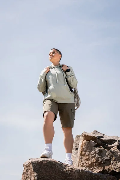 Low angle view of confident young short haired female tourist with fitness tracker and backpack standing on stones and looking away with blue sky at background, vibrant travel experiences — Stock Photo