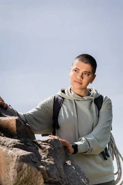 Jovem caminhante mulher de cabelos curtos com mochila olhando para longe enquanto estava perto de pedras e olhando para longe com o céu azul no fundo, experiências de viagem vibrantes, verão — Fotografia de Stock