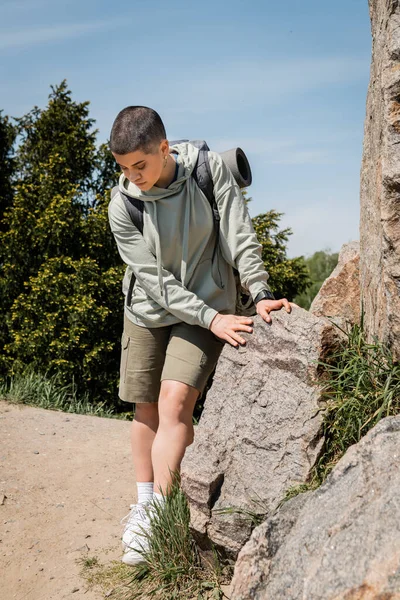 Young short haired female hiker in casual clothes with backpack looking at sneakers while standing near stones with trees and blue sky at background, trekking through rugged terrain — Stock Photo