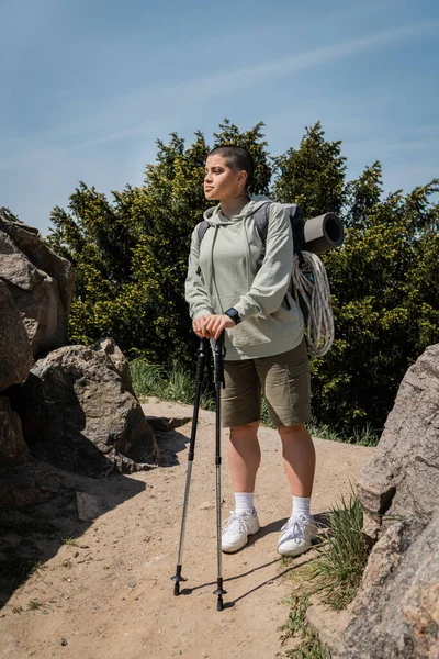 Young short haired and tattooed female hiker with fitness tracker and backpack with travel equipment holding trekking poles while standing on path with stones, trekking through rugged terrain — Stock Photo