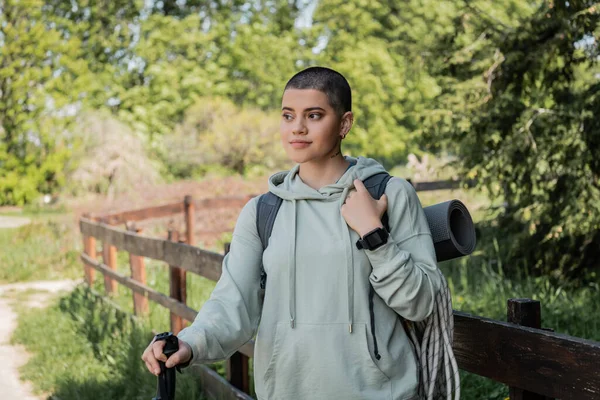 Young short haired woman hiker with backpack and fitness tracker holding trekking pole and looking away while standing with blurred landscape at background, trekking through rugged terrain — Stock Photo