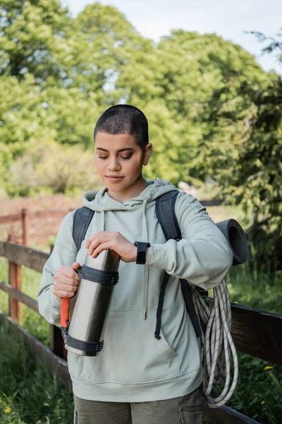 Young short haired female tourist with backpack and fitness tracker holding thermos while standing with blurred natural landscape at background, trekking through rugged terrain, summer — Stock Photo