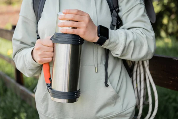 Cropped view of young woman tourist with backpack and fitness tracker holding thermos while hiking with blurred nature at background, trekking through rugged terrain, summer — Stock Photo
