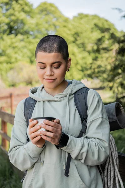Portrait of young short haired female traveler with backpack and travel equipment holding cup of thermos and standing with blurred landscape at background, finding serenity in nature — Stock Photo