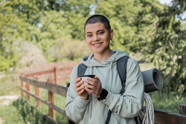 Веселый молодой коротковолосый женский турист с фитнес-трекером и рюкзаком держа термос чашку и глядя на камеру с пейзажем на заднем плане, находя спокойствие в природе, летом — стоковое фото