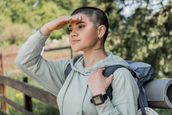 Young short haired and tattooed woman hiker with fitness tracker and backpack looking away while standing with blurred landscape at background, finding serenity in nature, summer — Stock Photo