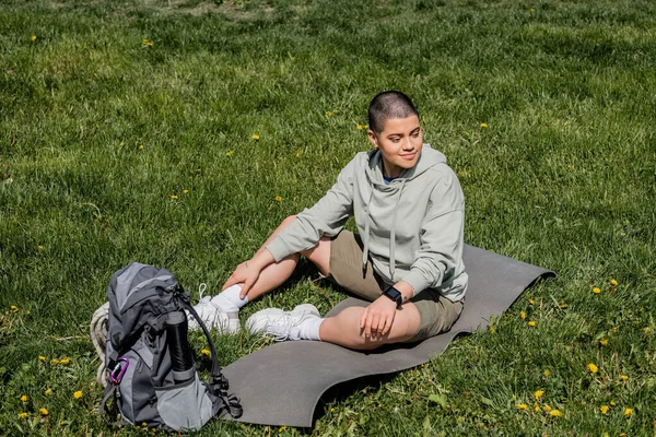 Smiling short haired female tourist sitting on fitness mat near backpack with travel equipment and looking away on grassy lawn with flowers, finding serenity in nature, summer — Stock Photo