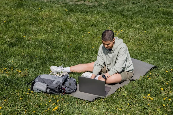 Junge kurzhaarige und tätowierte Touristin mit Laptop, während sie auf einer Fitnessmatte in der Nähe eines Rucksacks auf einer Rasenfläche mit Blumen sitzt, Ruhe in der Natur findet, Sommer — Stockfoto
