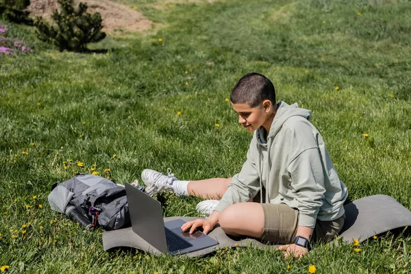 Jeune touriste aux cheveux courts en vêtements décontractés à l'aide d'un ordinateur portable assis sur un tapis de fitness près du sac à dos sur une pelouse herbeuse avec des fleurs, trouver la sérénité dans la nature, l'été — Photo de stock
