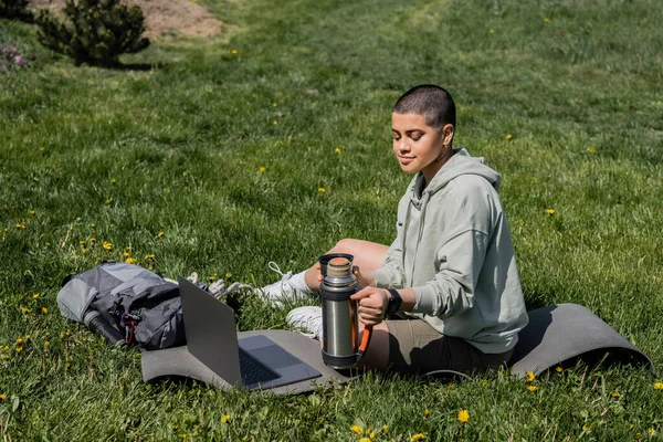 Junge kurzhaarige Touristin hält Thermoskanne in der Hand, während sie auf einer Fitnessmatte neben Laptop und Rucksack auf einer Rasenfläche mit Blumen sitzt, und findet Ruhe in der Natur, im Sommer, im digitalen Nomaden — Stockfoto