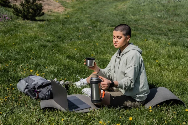 Junge tätowierte und kurzhaarige Wanderin mit Thermoskanne in der Hand, während sie neben Rucksack und Laptop auf einer Fitnessmatte auf dem Rasen mit Blumen sitzt und Gelassenheit in der Natur, im Sommer, im digitalen Nomaden findet — Stockfoto