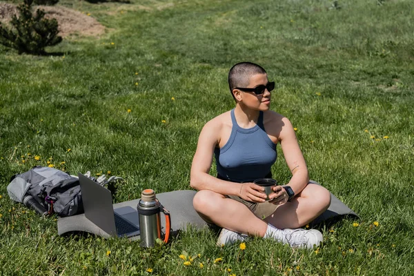 Young short haired female traveler in sunglasses holding thermos cup while sitting near laptop and backpack on fitness mat and lawn with flowers, finding serenity in nature, summer — Stock Photo