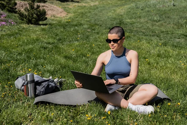 Молода короткошерста жінка-туристка в сонцезахисних окулярах використовує ноутбук, сидячи на фітнес-маті біля рюкзака і термос на трав'янистому газоні з квітами, знаходячи спокій в природі, літо, цифровий кочівник — стокове фото