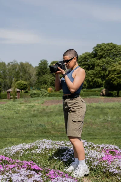 Short haired female tourist in sunglasses and fitness tracker taking photo on digital camera while standing on lawn with flowers with blurred nature at background, connecting with nature concept — Stock Photo