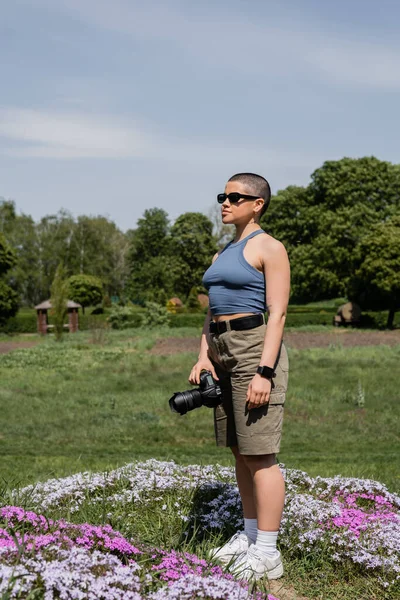 Jovem de cabelos curtos e tatuado viajante feminino em óculos de sol segurando câmera digital enquanto está em pé no gramado gramado gramado com flores e natureza turva no fundo, fotógrafo de viagens — Fotografia de Stock