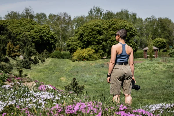 Vue latérale d'une jeune randonneuse aux cheveux courts et tatouée portant des lunettes de soleil tenant un appareil photo numérique debout sur la pelouse avec des fleurs et de la nature en arrière-plan, en connexion avec le concept de la nature — Photo de stock