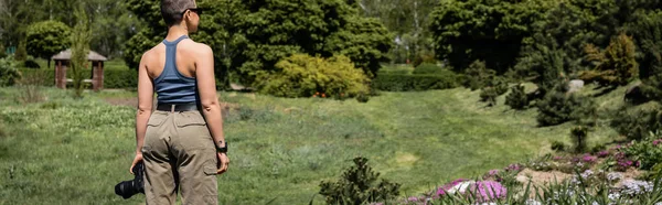 Seitenansicht einer jungen kurzhaarigen Touristin mit Sonnenbrille, die eine Digitalkamera hält und in der Nähe von Blumen steht, mit einer malerischen Landschaft im Hintergrund, die sich mit dem Naturkonzept verbindet, Banner — Stockfoto