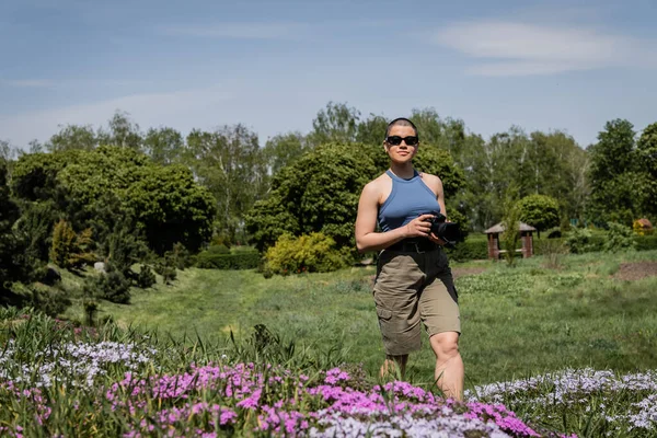 Молода короткошерста жінка-туристка в сонцезахисних окулярах тримає цифрову камеру, стоячи біля розмитих квітів з розмитим мальовничим пейзажем на фоні, переклад татуювання: любов — стокове фото