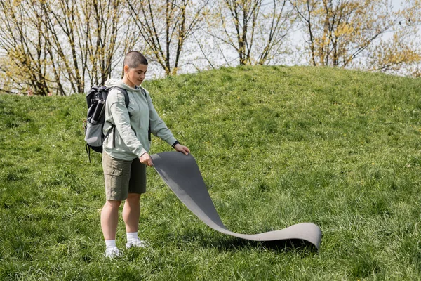 Sorridente giovane turista dai capelli corti femminile in abiti casual con zaino mettendo tappeto fitness su erba con collina e natura sullo sfondo, in collegamento con il concetto di natura, estate — Foto stock