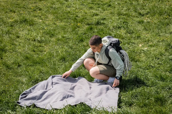 Hochblick auf junge kurzhaarige Wanderin mit Fitness-Tracker, Rucksack und Reiseausrüstung, die Decke auf den Rasen legt, Verbindung mit Naturkonzept, Sommer — Stockfoto