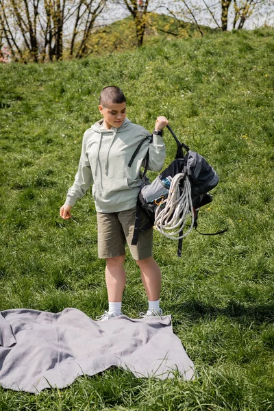 Jovem caminhante feminina de cabelos curtos com rastreador de fitness segurando mochila com equipamento de viagem enquanto está perto de cobertor no gramado gramado gramado com colina no fundo, conectando-se com o conceito de natureza — Fotografia de Stock