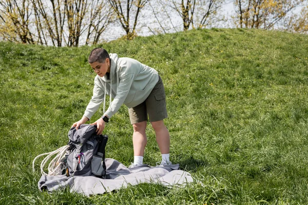 Молодая коротковолосая женщина-путешественница с фитнес-трекером положить рюкзак с туристическим оборудованием на одеяло, стоя на травянистой лужайке с холмом на заднем плане, подключаясь к концепции природы — стоковое фото