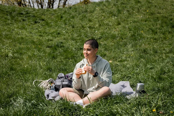 Веселая молодая коротковолосая и татуированная женщина-туристка держит сэндвич рядом с термосом и рюкзаком, сидя на травянистой лужайке с холмом на заднем плане, соединяясь с природой — стоковое фото