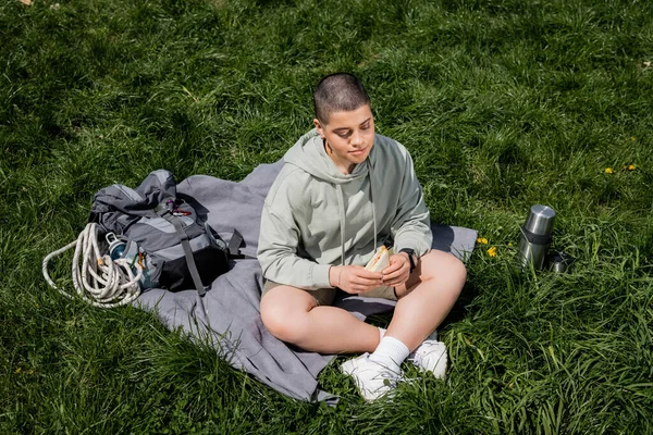 Vista de ángulo alto del joven senderista de pelo corto sosteniendo un sándwich mientras está sentado cerca de la mochila y el termo en la manta y el césped cubierto de hierba, conectando con el concepto de la naturaleza, Traducción del tatuaje: amor - foto de stock