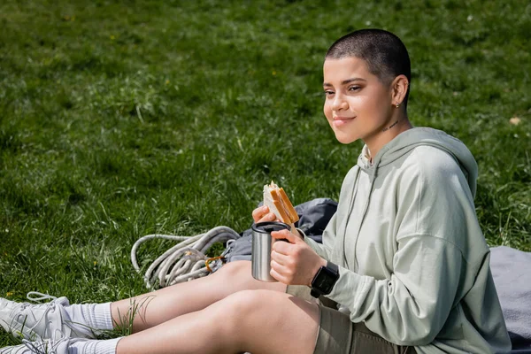 Smiling young short haired and tattooed female tourist holding sandwich and thermos cup while sitting near backpack on blanket on grassy lawn, connecting with nature concept — Stock Photo