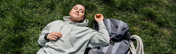 Top view of young relaxed and short haired female tourist with fitness tracker lying with closed eyes near backpack on grassy lawn, solo hiking journey concept, banner , summer — Stock Photo