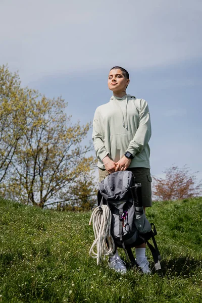 Молодая коротковолосая туристка с умными часами, держащая рюкзак с туристическим оборудованием и глядя в сторону, стоя на травянистом холме, пробираясь сквозь живописный пейзаж, лето — стоковое фото